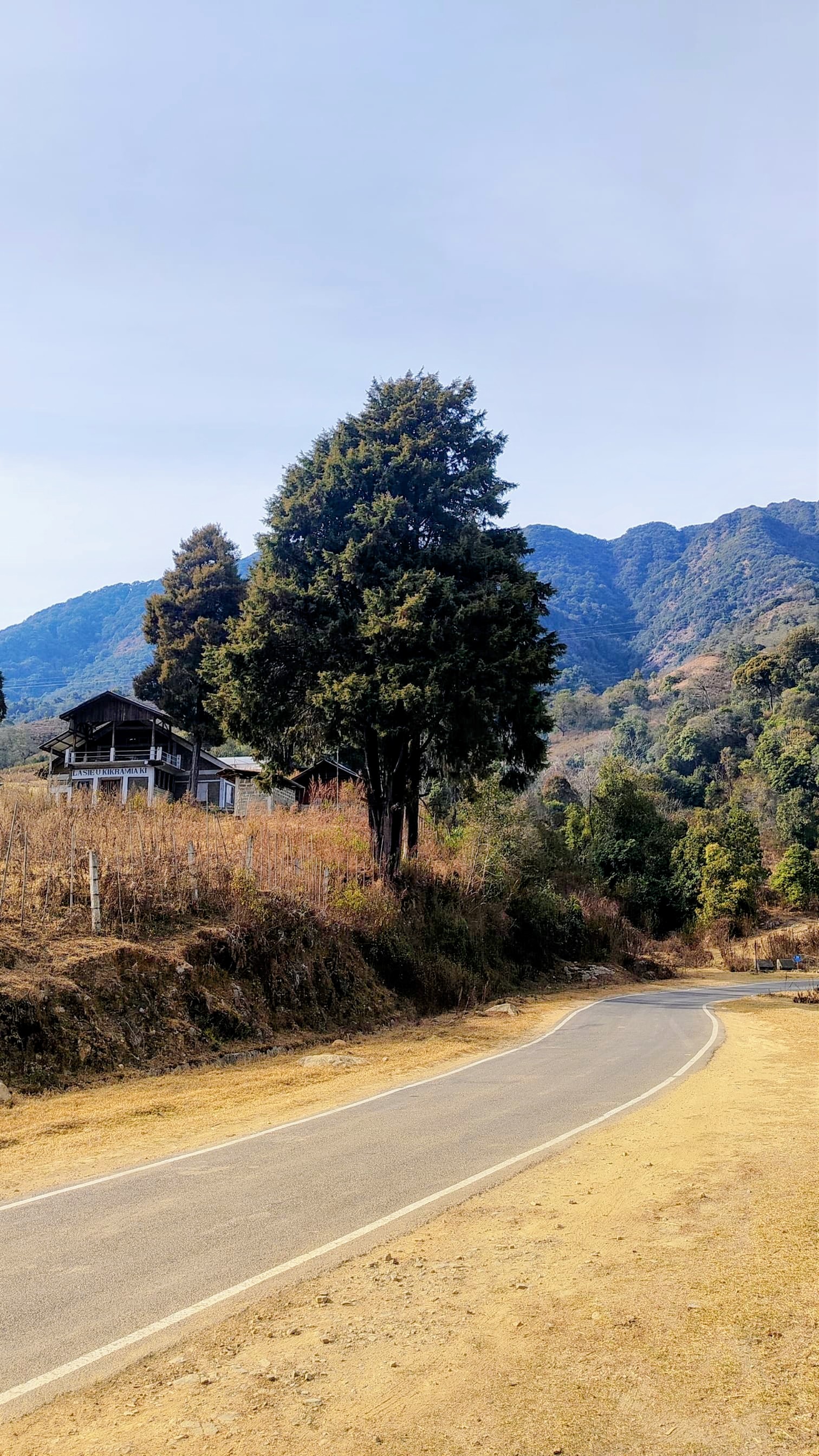 A quiet mountain road curving past a traditional wooden house with mountains in the distance